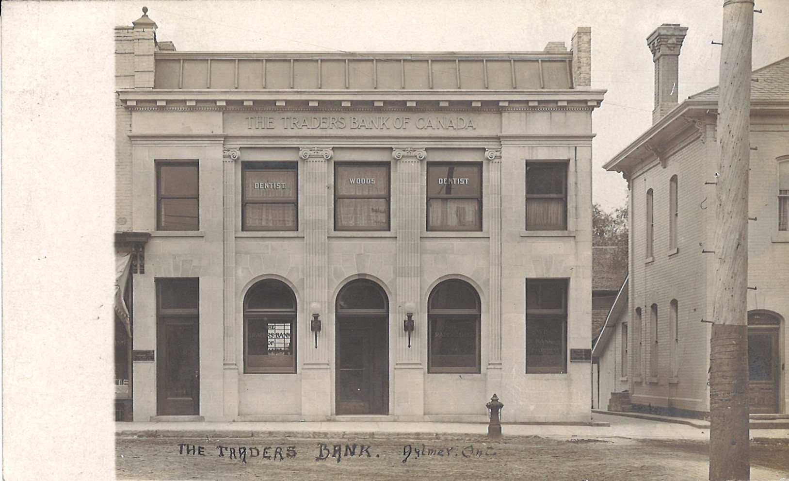 Photograph of The Traders Bank of Canada, circa 1909. The building, at the time, housed Dr. Woods, a dentist, in its upper level. The windows of the upper level read "DENTIST WOODS DENTIST". The façade of the building features four inset Ionic columns. The current façade, in comparison features four Doric columns, as the volutes of the columns' capitals are missing. 