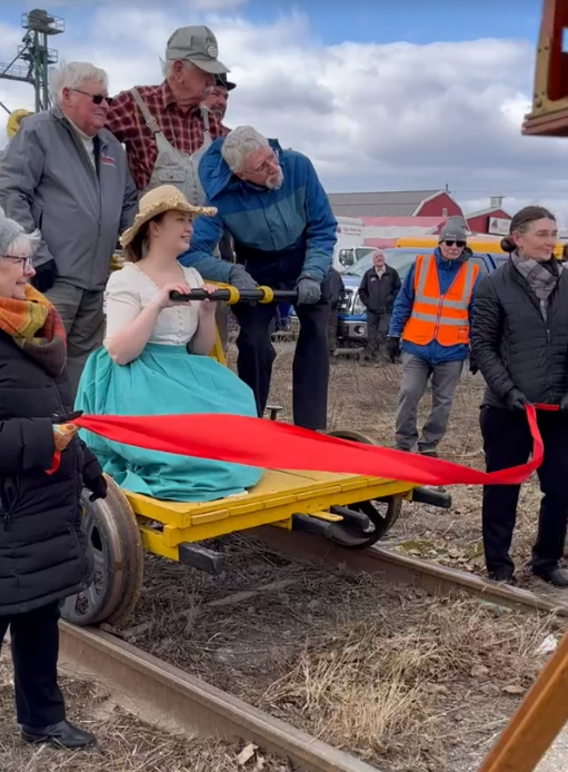 Museum representatives aboard the railcar alongside David Harding and Ed Ketchabaw. Betty Segui and Sarah Leitch hold a red ribbon in front of the handcar.