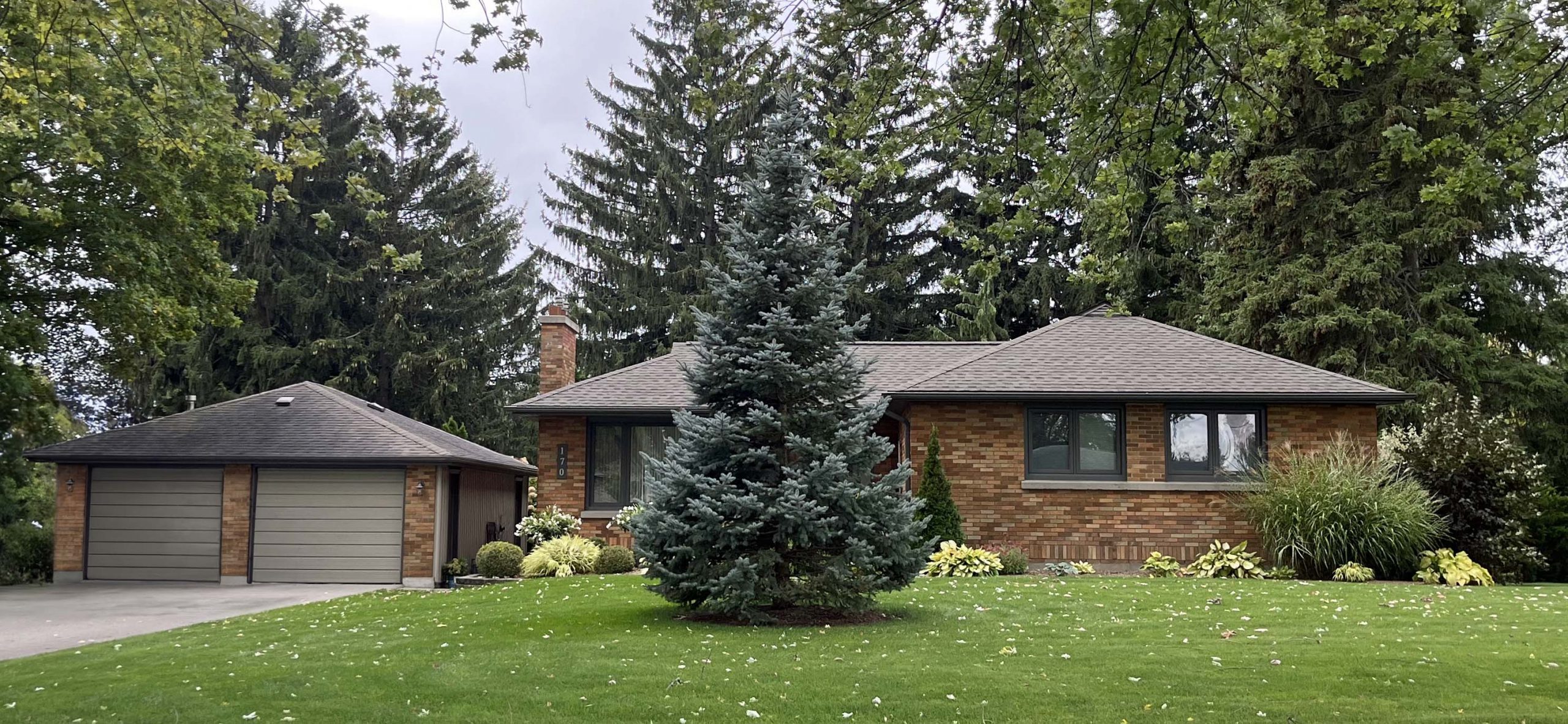 8394 Caverly Rd. A single-storey red-brown brick home with a double-car garage. partially obscured by and surrounded by pine trees.