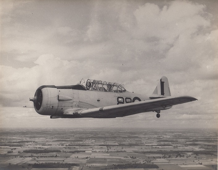 Image of a plane, mid-flight, with its pilot and passenger visible within, and farmland below.
